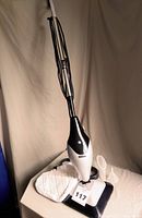 Full view of the Bionaire steam mop standing upright with visible handle and body, placed next to cleaning pads and accessories on a beige surface with a neutral background.