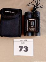 Photo showing Tasco binoculars folded and placed next to a black Tasco branded carry case, Lot #73 indicated.