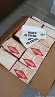 Box containing multiple white paper caps stacked and several boxes labeled 'Gellwrap'. Caps have 'HOOD'S ICE CREAM 25¢' branding.