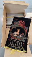 Opened box showing dozens of rolled vintage Coca Cola 1899 calendar tapestries in black felt with printed design and yellow fringe.