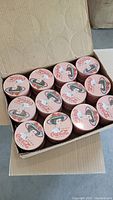 Box containing 12 Coca-Cola vintage tins with pink background and a woman's face wearing a rose hat on the lid.