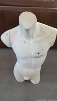 Front view of a white male torso mannequin showing muscular chest and visible wear marks, on a floor surface.