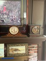 Two antique mantle clocks displayed side by side on a mantelpiece beneath framed artwork. Left is Seth Thomas tambour style clock and right is Sessions Clock Co mantle clock with rectangular wooden case.