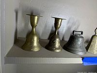 Two brass candleholder-style bells, one metal handle bell, and one smaller metal bell with loop handle on a shelf.
