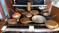 Photo of all copper tone cookware and accessories arranged on a stove, including pots with lids, large colander, container with ceramic handles, bed warmer with wooden handle, and various pans.