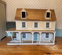 Front view of wooden dollhouse with beige siding and brown shingled roof, featuring porch and turquoise door
