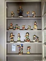Photo showing 14 figurines on three shelves inside a cabinet, mostly children figurines with various poses and items