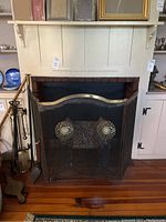 Front view of black metal fireplace screen with decorative elements, placed in fireplace with andirons beside it.