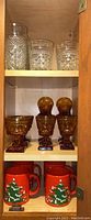 Four red Waechtersbach Christmas mugs with green tree decoration on bottom shelf, and four amber stemware glasses on middle shelf