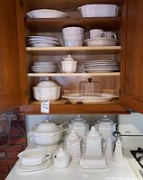 Front view of cupboard shelves with stacked plates, bowls, cups, and covered serving dishes.