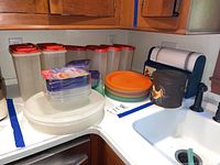 Kitchen corner counter showing plastic storage containers with red lids, colorful plates, paper towel holder, and tin container