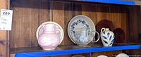 Six items arranged on a wooden shelf including glass teapot, two china plates, pink floral pitcher, wooden chargers, glazed clay pot with blue decoration