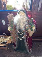 Full view of the Santa Claus figure holding twig branches and a burlap sack with greenery, placed near a window with natural light. Visible red robe with fur trim and curly white beard.