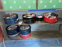 Eight decorative tins on a wooden shelf, shown from the front to display their lids and contents partially visible.