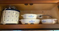 Set of three white Corning Ware casserole dishes with blue floral pattern stacked, alongside a Corning Ware slow cooker with matching blue floral tile design on a wooden shelf.