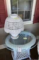 Photo showing a white wicker side table with a round removable glass top and a matching lamp with wicker shade on top.