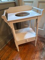 Front angle image of white wooden washstand with basin cutout, drawer, and shelf on wooden floor by window.