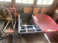 Photo showing a black plastic log carrier next to a red metal-handled wheelbarrow in a garage with wooden walls and concrete floor.