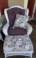 Front view of white wicker chair with tufted dark brown cushions and floral throw pillow next to matching ottoman with floral cushion, showcasing chair and ottoman design and condition.
