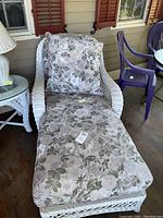 Full view of white wicker chaise lounge with floral cushion on porch alongside matching side table and plastic chairs.