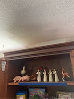 Shelf display showing 4 Willow Tree figurines alongside 3 animal figurines including a ceramic bird with red head and a light pink rabbit figurine lying down. Wooden cabinet and children's educational materials below shelf.