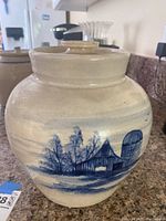 Front view showing the blue painted rural scene on white ceramic pottery jar with lid.