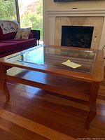 Photo showing the coffee table with glass top, wood frame and carved legs, placed on wood floor in front of fireplace and couch.