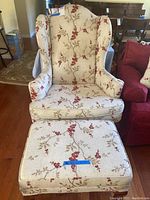 High back floral print wingback chair with matching ottoman, showing fabric pattern and wooden legs, placed on hardwood floor beside red chair and dining table in background.