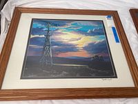 Framed signed print showing wooden windmill against dramatic sunset sky with clouds, matted and in wood frame