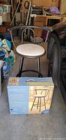 Photo showing one used black metal swivel bar stool with a light-colored microfiber seat cushion and the unopened box containing the other new bar stool.