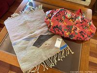 Photo showing Navajo style woven rug partially unfolded with black geometric pattern and fringe; floral patterned red oil cloth tote bag next to it on a glass table.