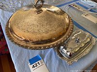 Photo showing large round silver-plated serving dish with engraved floral patterns and lid with handle, alongside small silver-plated butter dish with ornate edges and lid.