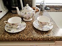 Photo of a white porcelain China tea set with purple and green floral design, includes one teapot with lid, two teacups, and two saucers arranged on a countertop.