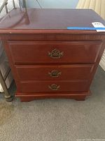 Front view of wooden nightstand showing three drawers with brass handles, reddish-brown finish, sitting on carpeted floor beside metal bedframe.