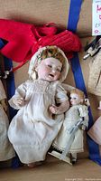 Two antique porcelain dolls placed side by side, large doll with red knitted clothing and white dress, smaller doll in white dress with bow.