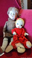 Two vintage dolls seated on red chair: porcelain boy doll in brown suit with damaged hands and feet, and fabric doll in red riding hood outfit.