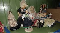 Seven antique dolls displayed together on a shelf with green wooden background, showing a variety of traditional costumes and materials.