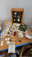 Wide view of lot items on table including display case, jewelry, boxes
