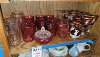 Overall shelf view of vintage glassware including colored pink and brown glasses, candy dish, stemware and decorative oil bottle.