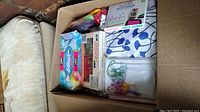 Photo of cardboard box containing Dot Snappers kit, blue floral fabric, note cards, and various small craft supplies including buttons, zippers, and ribbons.