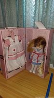 Doll wearing blue dress holding a stuffed bunny inside an open pink wardrobe-style case with visible hanging doll clothes.