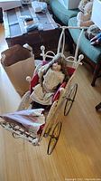 Wide view of porcelain doll seated in vintage white wicker stroller with metal spoked wheels on hardwood floor.
