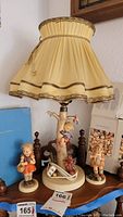 Lamp with fabric shade featuring Hummel figurine base and two other Hummel figurines on wooden surface