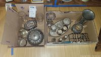 Wide angle photo showing the total lot in two cardboard boxes, containing various pewter and plated servingware including pitchers, salt and pepper shakers, dishes, glasses, and vases.