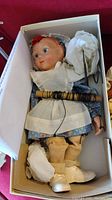 Marionette doll lying in box, showing seated pose with strings and protective paper around feet and hands.
