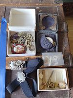 Lot contents arranged on a wooden chest including pocket watch in case, costume jewelry in white boxes, and black fabric belt with metal buckles.