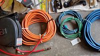 Overview of orange retractable hose reel, orange hose, blue and green extension cords and hoses on garage floor.