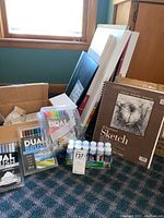 Overview of art supplies including paint bottles, sketch pads, pens, and canvases on carpeted floor near window.