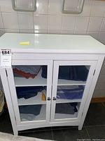 White wooden cabinet with two glass-paneled doors, placed against tiled wall, showing the overall item and contents inside.