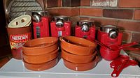 Four red square metal dry goods cannisters with silver lids, six terracotta round fondue baking dishes stacked, one orange-brown Nescafe canister with cream lid, red measuring spoons and cups